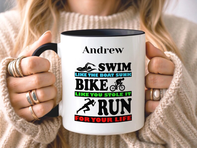 May include: White ceramic mug with a black interior and handle. The mug features the name "Andrew" above the words "SWIM, BIKE, RUN" with related graphics and phrases. The mug is held by a person wearing rings.