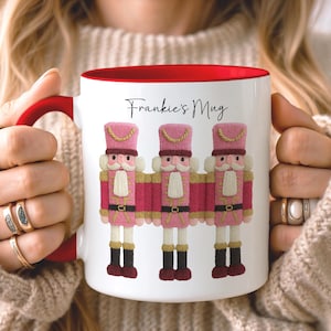 May include: White ceramic mug with a red interior and handle, featuring three pink and red nutcracker figures. The mug has the text "Frankie's Mug" in black script. The design is festive and whimsical.