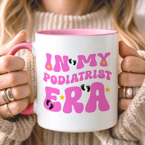 May include: A white ceramic mug with a pink handle and interior rim. The front features the pink text "IN MY PODIATRIST ERA" in a bold, wavy font, accented with small black footprints and yellow stars scattered throughout the lettering.