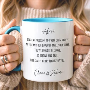 Op de afbeelding: Een witte keramische mok met een felblauw handvat en binnenrand. De mok is voorzien van zwarte tekst: Alex, Today we welcome you with open hearts, As you and our daughter make your start. You've brought her love, So strong and true, Our family grows because of you. Clare & Zak xx.