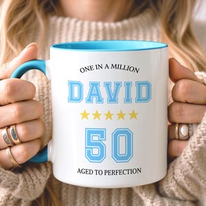 May include: A white ceramic mug with a bright blue interior and handle. The front features the text ONE IN A MILLION, the name DAVID in blue block letters, five yellow stars, the number 50 in blue, and the text AGED TO PERFECTION.