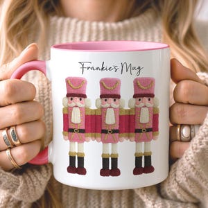 May include: White ceramic mug with a pink interior and handle, featuring three pink and gold nutcracker figures. The mug has the text "Frankie's Mug" in black script.