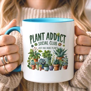 May include: A white ceramic mug with a bright blue handle and interior rim. The front features a colorful graphic of various potted succulents and cacti with the text "PLANT ADDICT SOCIAL CLUB" and "JUST ONE MORE PLANT" printed in green.