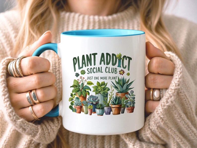 May include: A white ceramic mug with a bright blue handle and interior rim. The front features a colorful graphic of various potted succulents and cacti with the text "PLANT ADDICT SOCIAL CLUB" and "JUST ONE MORE PLANT" printed in green.