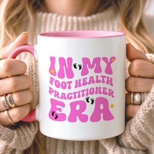 May include: White ceramic mug with a pink interior and handle. The mug features the text "IN MY FOOT HEALTH PRACTITIONER ERA" in pink, with star and footprint graphics. The mug is held by a person.
