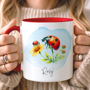 Puede incluir: Taza de cerámica blanca con interior y asa rojos. La taza presenta una ilustración de acuarela de dos mariquitas sobre una flor amarilla con un fondo azul. El nombre "Rosy" está escrito en cursiva.