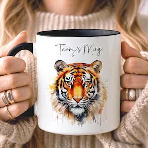 Könnte beinhalten: Ein weißer Keramikbecher mit schwarzem Henkel und schwarzem Innenrand. Auf der Vorderseite befindet sich eine farbenfrohe Aquarell-Illustration eines Tigergesichts unter dem schwarzen, kursiven Schriftzug Terry's Mug auf der glatten Oberfläche.