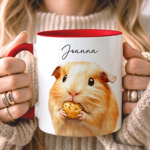 May include: White ceramic mug with a red interior and handle, featuring a watercolor illustration of a guinea pig holding a snack. The name "Joanna" is written in black script. A charming and personalized gift.