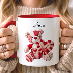 Pode incluir: Caneca de cerâmica branca com interior e alça vermelhos, com um urso de pelúcia de retalhos segurando um coração. O urso é vermelho, branco e rosa com estampas florais e xadrez. O nome "Freya" está escrito em itálico preto.