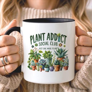 May include: A white ceramic mug with a black handle and rim, featuring a colorful graphic of various potted succulents and plants. The text reads "PLANT ADDICT SOCIAL CLUB" in green letters above the phrase "JUST ONE MORE PLANT" in smaller black text.