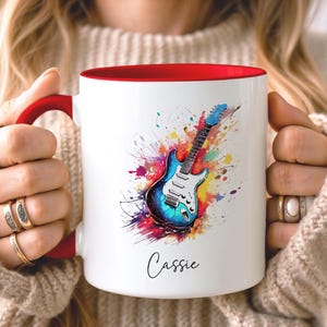 Puede incluir: Una taza de cerámica blanca con interior y asa rojos, que presenta una colorida ilustración estilo acuarela de una guitarra eléctrica rodeada de salpicaduras de pintura vibrantes. El nombre Cassie está impreso en negro bajo el gráfico.
