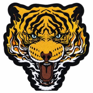 Puede incluir: Un parche bordado que muestra la cabeza de un tigre con pelaje amarillo, rayas negras y ojos azul claro. El tigre está rodeado de llamas estilizadas de color naranja, amarillo y blanco, todo delineado con un borde negro grueso y boca marrón.