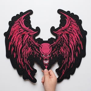 May include: A large embroidered patch featuring a central demonic figure with sharp teeth and glowing white eyes, flanked by two expansive, jagged wings. The design is rendered in vibrant hot pink thread against a solid black background.