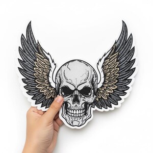 May include: A detailed patch featuring a skull with feathered wings. The skull is gray with black details, and the wings are gray and tan. The patch has a white border and is held against a white background.