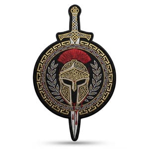 Puede incluir: Un parche bordado que presenta un casco espartano central en dorado y plateado con una cresta roja, colocado detrás de una espada vertical. El diseño está enmarcado por un borde circular con un patrón de greca dorada y hojas de laurel.