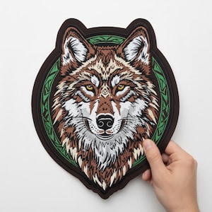 May include: Embroidered patch featuring a detailed wolf head design. The patch has a brown border and a green circular accent with a Celtic knot pattern. The wolf's fur is brown, white, and tan, with yellow eyes.
