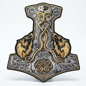 May include: An embroidered patch shaped like Mjolnir, featuring a central face with a long beard, two howling wolves in gold thread, and two smaller animals at the base. The design incorporates intricate silver and black knotwork patterns.