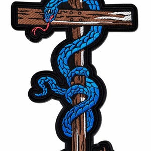 May include: An embroidered patch featuring a bright blue snake with a red tongue coiled around a brown wooden cross. The cross has a textured appearance with white highlights, and the entire design is outlined with a thick black border.