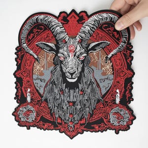 May include: Embroidered patch featuring a detailed illustration of a goat with large horns, red eyes, and intricate patterns. The patch is primarily black, red, and gray, with a heart-shaped border and decorative elements. The goat has long, flowing hair.
