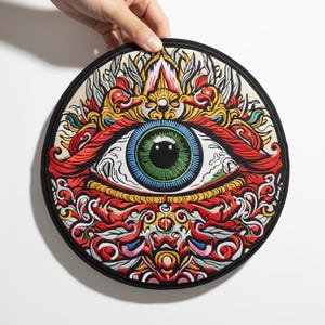 May include: A round, embroidered patch featuring a detailed eye design. The eye has a green iris, blue and white details, and is surrounded by ornate red, yellow, and gold embroidery. The patch has a black border and is held by a hand.