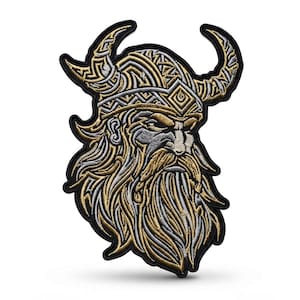 Può includere: Una toppa ricamata raffigurante la testa di un guerriero vichingo con un elmo cornuto. Il design utilizza filo metallico dorato e argentato su sfondo nero, mostrando intricati motivi a intreccio sull'elmo e una barba fluente e dettagliata.
