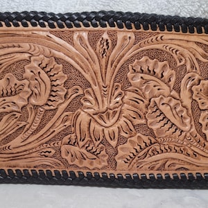 Könnte beinhalten: Eine rechteckige, hellbraune Lederbrieftasche mit detailliertem Blumendesign. Das Design ist in das Leder geprägt, mit einem schwarzen geflochtenen Rand. Die Brieftasche hat ein natürliches, rustikales Aussehen.