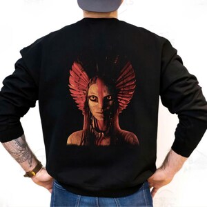 Pode incluir: Camisola preta com um gráfico marcante nas costas. O design apresenta uma pessoa com pintura facial vermelha e branca e um cocar de penas. A parte superior de manga comprida é uma peça ousada.