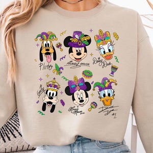 Op de afbeelding: Beige sweatshirt met kleurrijke cartoon illustraties van Disney-personages Pluto, Mickey Mouse, Daisy Duck, Goofy, Minnie Mouse en Donald Duck, allemaal in Mardi Gras-outfits. De namen van de personages staan eronder geschreven.