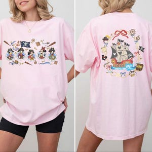 Puede incluir: Camiseta rosa claro con un diseño de temática pirata. La parte delantera muestra personajes de dibujos animados vestidos de piratas, mientras que la parte trasera presenta una ilustración de un barco pirata rodeado de elementos náuticos.