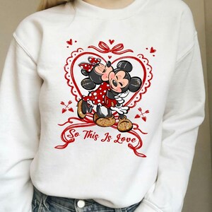 May include: White sweatshirt featuring a graphic of Mickey and Minnie Mouse kissing inside a red heart. Minnie wears a red polka dot dress and bow. The text "So This Is Love" is written in red script.