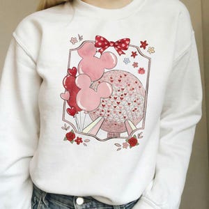 May include: White sweatshirt with a graphic design. The design includes pink Mickey Mouse balloons, red heart balloons, and a red and pink dotted bow. A pink and red sphere with heart details and floral accents is also featured.