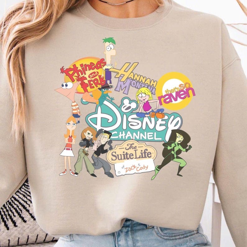 Pode incluir: Camisola bege com um gr&aacute;fico colorido de personagens do Disney Channel, incluindo Phineas e Ferb, Hannah Montana e Kim Possible. O log&oacute;tipo do Disney Channel &eacute; vis&iacute;vel.