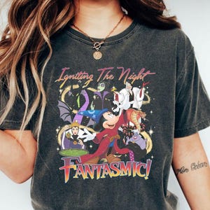 Puede incluir: Camiseta gris oscuro con un gr&aacute;fico colorido de Mickey Mouse y otros personajes de Disney. El texto "Igniting The Night" y "Fantasmic!" se muestran en una fuente estilizada.