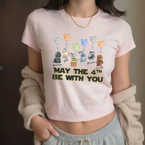 May include: A pale pink t-shirt with a graphic of Star Wars characters as balloons with Mickey Mouse ears. The text "MAY THE 4TH BE WITH YOU" is printed in yellow and black. The shirt is a cropped style.