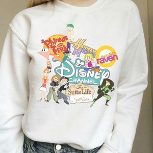 Pode incluir: Camisola branca com gr&aacute;ficos coloridos de desenhos animados de programas do Disney Channel, incluindo "Phineas e Ferb", "Hannah Montana", "As Vis&otilde;es da Raven" e "Zack & Cody: G&ecirc;meos em A&ccedil;&atilde;o". O design &eacute; uma colagem de personagens e logotipos.