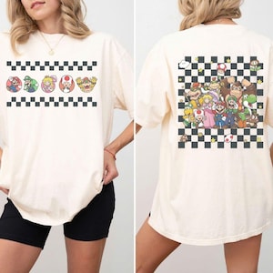 以下が含まれることがあります： クリーム色のTシャツ。マリオのキャラクターのカラフルなグラフィックが特徴です。前面にはキャラクターの肖像画が入ったチェック柄のボーダーがあります。背面には、チェック柄の背景にキャラクターの大きなグループシーンが表示されています。楽しくてカジュアルなTシャツです。