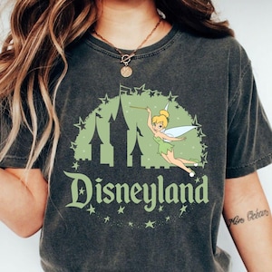 May include: Dark grey crew neck t-shirt with a green Disneyland graphic. The design includes a castle, stars, and Tinkerbell. The text "Disneyland" is in a decorative font. The shirt is a casual wear item.