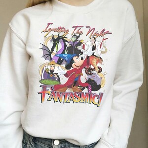 Puede incluir: Sudadera blanca con un gr&aacute;fico colorido de personajes de Disney, incluyendo a Mickey Mouse y villanos. El dise&ntilde;o presenta el texto "Igniting The Night Fantasmic!". El gr&aacute;fico incluye un drag&oacute;n y una serpiente.