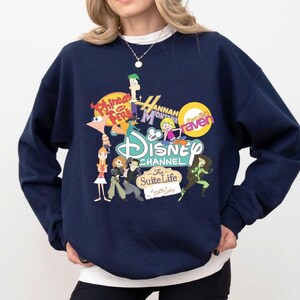 Pode incluir: Camisola azul marinho com um gr&aacute;fico colorido do Disney Channel. O gr&aacute;fico inclui personagens e logotipos de v&aacute;rios programas da Disney, como Phineas e Ferb, Hannah Montana e Kim Possible. A camisola tem gola redonda.