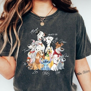 May include: Dark gray t-shirt featuring a colorful illustration of various cartoon dogs, including Pongo, Perdita, and Pluto. The names of the dogs are written above them in a handwritten style. The shirt has a crew neck and short sleeves.