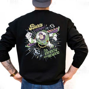 Puede incluir: Sudadera negra con un diseño de Buzz Lightyear en su traje espacial. El diseño incluye el texto "Buzz Lightyear" y "To Infinity and Beyond" en un estilo retro, con detalles en verde, morado y blanco.