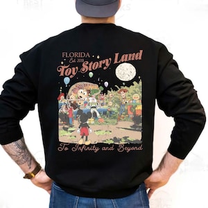 Puede incluir: Sudadera negra con un gráfico colorido que representa una escena de parque temático con el texto "Toy Story Land" y "To Infinity and Beyond". El diseño incluye personajes de dibujos animados y las palabras "Florida Est. 2018."