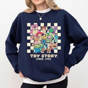 May include: Navy blue sweatshirt featuring a graphic of Toy Story characters on a checkered background. The text "TOY STORY. SINCE 1995" is printed below the characters. The sweatshirt has a crew neck and long sleeves.