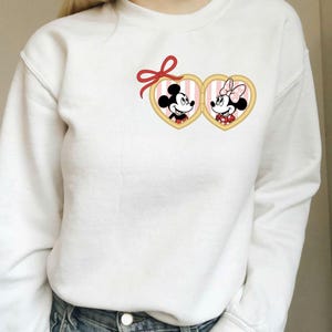 Op de afbeelding: Witte sweatshirt met een Mickey en Minnie Mouse ontwerp. Het ontwerp toont Mickey en Minnie in hartvormige frames met roze en witte strepen, bekroond met een rode strik. Een casual, comfortabele stijl van sweatshirt.