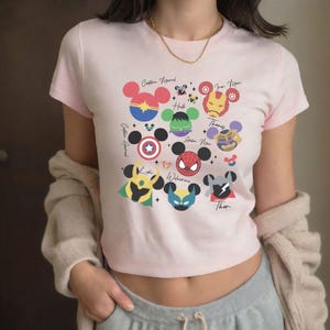 Pode incluir: Um top cropped rosa claro com um design gráfico de vários personagens da Marvel estilizados como cabeças do Mickey Mouse. Os personagens incluem Capitã Marvel, Hulk, Homem de Ferro, Capitão América, Homem-Aranha, Wolverine e Thor. Os nomes são impressos.