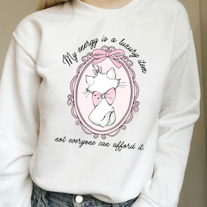 Puede incluir: Sudadera blanca con una ilustración de gato de dibujos animados dentro de un marco ovalado rosa. El gato lleva un lazo rosa y el texto dice: "My energy is a luxury item, not everyone can afford it."