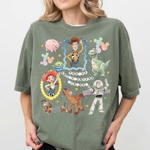 Può includere: T-shirt verde salvia con personaggi dei cartoni animati del film Toy Story. Il design include Woody, Buzz Lightyear, Jessie, Rex, Hamm e altri personaggi. È presente anche il testo "You've Got a Friend in Me".