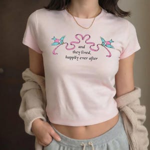 May include: A light pink t-shirt featuring two blue birds with pink accents and a pink ribbon design. The text "and they lived, happily ever after" is printed below the birds. The shirt has short sleeves.