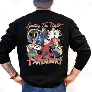 Puede incluir: Sudadera negra con un gr&aacute;fico colorido de personajes de Disney, incluyendo Mickey Mouse, y villanos. El texto "Igniting The Night" y "FANTASMIC!" se muestran en una fuente vibrante y multicolor.