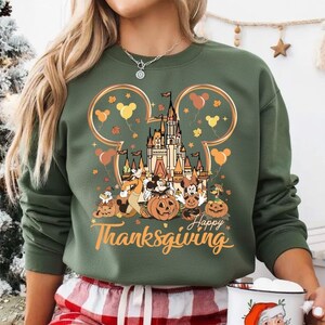 Op de afbeelding: Groen sweatshirt met een Disney-thema Thanksgiving-afbeelding. Het ontwerp bevat een kasteel, Mickey en Minnie Mouse, pompoenen en ballonnen. De woorden "Happy Thanksgiving" staan in oranje letters. De sweatshirt heeft een ronde hals.